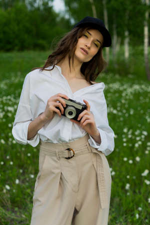 Beautiful girl photographer wearing a white shirt, beige trousers poses outdoors with a vintage camera against a background of meadow full of wild flowers. Lifestyle and commercial design. Copy space.の写真素材