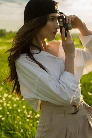 Beautiful girl photographer wearing a white shirt, beige trousers poses outdoors with a vintage camera against a background of meadow full of flowers in sunset light. Lifestyle design. Copy space.の写真素材