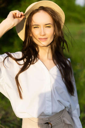 Beautiful stylish young girl wearing a trendy white shirt, beige trousers and boater hat smiling poses outdoors against a background of green leaves. Advertising and fashion design. Copy space.の写真素材