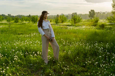 Beautiful girl photographer wearing a white shirt, beige trousers poses outdoors with a vintage camera against a background of meadow full of flowers in sunset light. Lifestyle design. Copy space.の写真素材