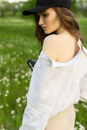 Beautiful girl photographer wearing a white shirt, beige trousers poses outdoors with a vintage camera against a background of meadow full of wild flowers. Lifestyle and commercial design. Copy space.の写真素材