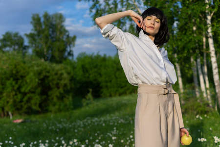 Beautiful stylish girl wearing a trendy white shirt, beige trousers and a black wig holds a green apple in sunlight on a background of a blue cloudy sky. Advertising and fashion design. Copy space.の写真素材