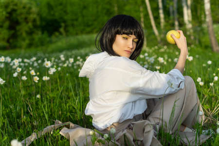 Beautiful stylish girl wearing a trendy white shirt, beige trousers and a black wig holds a green apple in sunlight sitting on a green meadow grass. Advertising and fashion design. Copy space.の写真素材