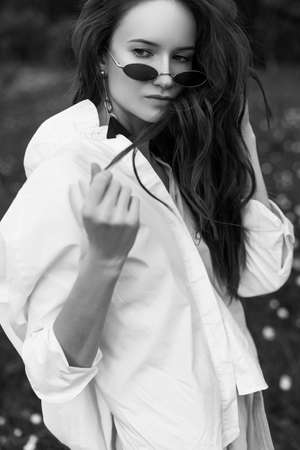 Art style black and white photo of a young stylish girl wearing a trendy white shirt, sunglasses outdoors on the meadow full of white wild flowers. Commercial and fashionable design. Copy spaceの写真素材