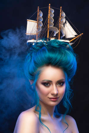 Sensual young girl with naked shoulders and painted blue hair stowed in an artistic hairdo with a toy sailboat in her hair, covering her chest with blue and white veils in a scenic smoke. Copy spaceの写真素材