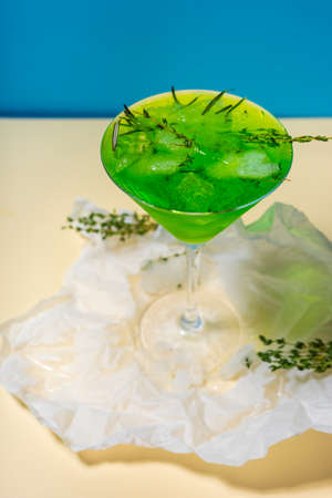 A green drink with ice cubes and green branches in a glass for mojito on crumpled wrapping paper on yellow table and blue background. Conceptual and advertising design. Copy space.の写真素材