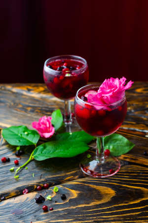 Two glasses full of red drink with berries, ice and a pink flowers, beside which lie berries and a branch of green tropical leaves on a vintage wooden table. Copy space. Commercial design.の写真素材