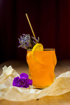 Figured glass of yellow fruit juice with a slice of lemon, a green branch of small flowers and a...