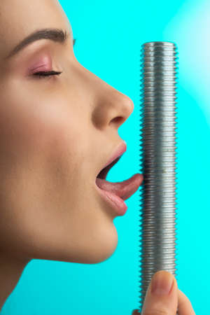 A beautiful girl with naked shoulders and closed eyes touches the thread of big steel bolt with her tongue. Close up. Isolated on blue. Copy space. Advertising and conceptual design. Healthy skin.の写真素材