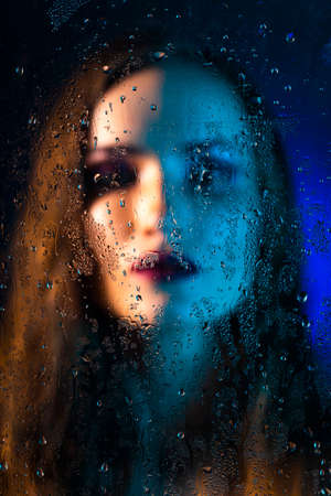 Art style portrait in blue tones of a lonely beautiful sad model girl behind a window glass, over which rain drops down. Artistic blur on model face. Conceptual, commercial and fashionable. Copy spaceの写真素材