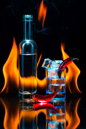 Blue shot glass full of ice, little bottle, two pods of red chile pepper and fire flames on a black background. Conceptual, commercial and advertising photo. Copy space.の写真素材