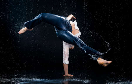 A young barefoot man wearing wet blue jeans and a white shirt expressively dances modern dances on the water under rainy waterdrops among the watersplashes. Isolated on a black background. Copy space.の写真素材