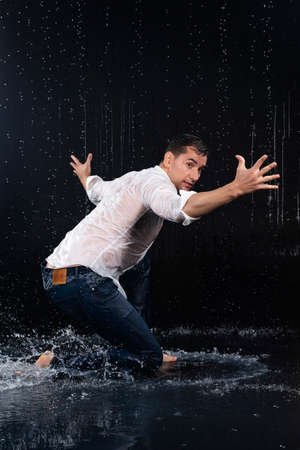 A young barefoot man wearing wet blue jeans and a white shirt expressively dances modern dances on the water under rainy waterdrops among the watersplashes. Isolated on a black background. Copy space.の写真素材