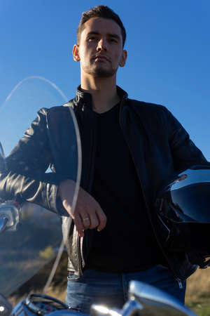 Young man wearing a black leather jacket holds mirror helmet in his hands and rests stays outdoor near a motorcycle. Lifestyle, travel. Copy space. Advertising and commercial design.の写真素材