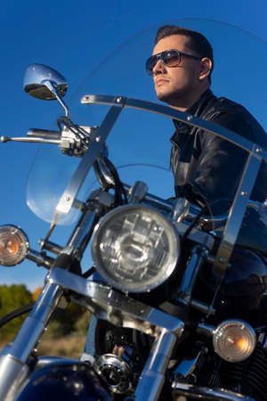 Young man wearing a black leather jacket, sunglasses and jeans sits outdoor on a motorcycle, on a mountain on a blue sky background. Lifestyle, travel. Copy space. Advertising and commercial design.の写真素材