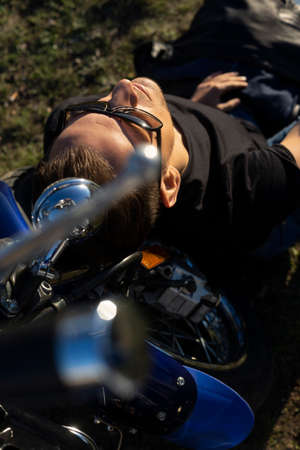 Young man wearing a black leather jacket, sunglasses and jeans sits outdoor on a ground at a motorcycle, resting on a mountain above the river. Lifestyle, travel. Copy space. Advertising design.の写真素材