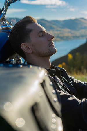 Young man wearing a black leather jacket and jeans sits outdoor on a ground at a motorcycle, resting...