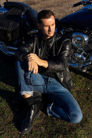 Young man wearing a black leather jacket and jeans sits outdoor on a ground at a motorcycle, resting...