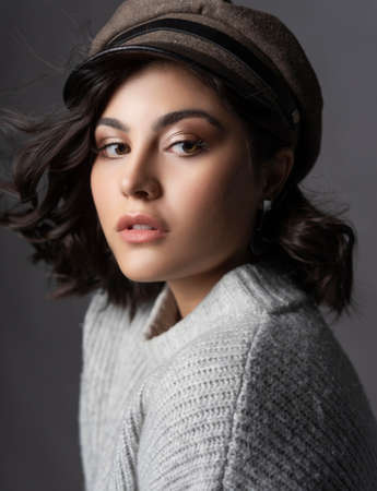 Closeup portrait of a beautiful brunette girl wearing a casual style knitted sweater and a cap on gray background. Natural nude makeup. Fashion, advertising and commercial design. Copy space.の写真素材