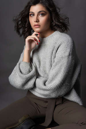 Beautiful brunette girl wearing casual style knitted sweater, brown pants, sits on a stool on a gray backgroundの写真素材