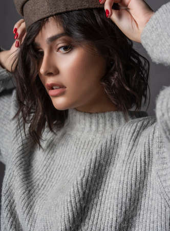 Beautiful scared brunette girl wearing a casual style knitted sweaterputs on cap on her head on gray background. Natural nude makeup. Fashion, advertising design. Close up. Copy spaceの写真素材