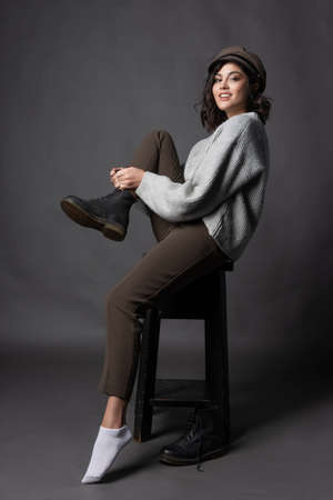 Beautiful smiling brunette girl wearing a casual style sweater, pants and cap puts a shoe on her leg on a gray background. Natural nude makeup. Fashion, advertising and commercial design. Copy space.の写真素材