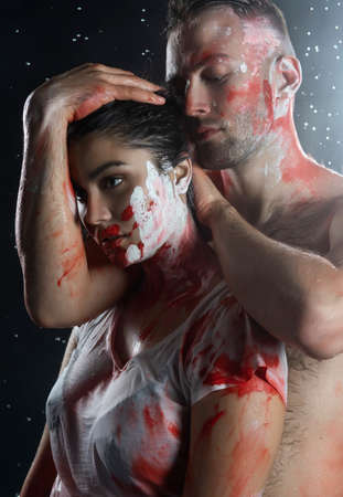 Couple of artists wearing jeans and t-shirts soiled with paint hug each other under rain in smoke on black background. Palm prints and paints on their faces. Man with naked torso. Copy spaceの写真素材