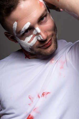 Stylish athletic male artist wearing a white t-shirt stained with red paint posing on gray background. White handprint of the palm on his face. Conceptual, fashion design. Copy space. Close up.の写真素材