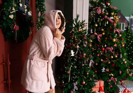 Beautiful smiling girl wearing a warm fluffy robe, wearing a hood over her head, stands at the red door decorated with fir branches and a Christmas tree. New year, xmas, lifestyle design. Copy space.の写真素材