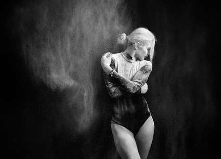 Beautiful slim girl wearing a gymnastic bodysuit covered with clouds of the flying white powder dances on a dark background. Artistic conceptual and advertising black and white photo. Copy spaceの写真素材