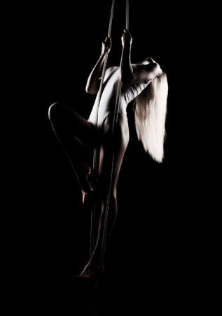 Beautiful slender blonde girl, wearing a white bodisuit and red stilettos, sensually hangs and dances on the ropes. Black background. Artistic noir silhouette photo. Copy space.の写真素材