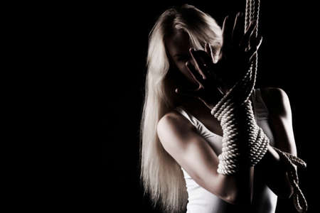 Beautiful slender blonde girl with big breast and nipples appearing through clothes, wearing a white bodisuit, sensually plays with the ropes on black. Artistic noir silhouette photo. Copy space.の写真素材
