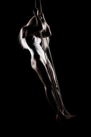 Beautiful slender blonde girl, wearing a white bodisuit and red stilettos, sensually hangs and dances on the ropes. Black background. Artistic noir silhouette photo. Copy space.の写真素材