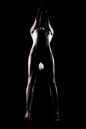 Beautiful slender blonde girl, wearing a white bodisuit and red stilettos, sensually hangs and dances on the ropes. Black background. Artistic noir silhouette photo. Copy space.の写真素材