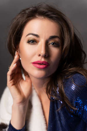 Closeup portrait of a beautiful brunette woman wearing a blue shiny dress and fur coat on a gray background. Clean, healthy skin. Advertising and commercial design.の写真素材