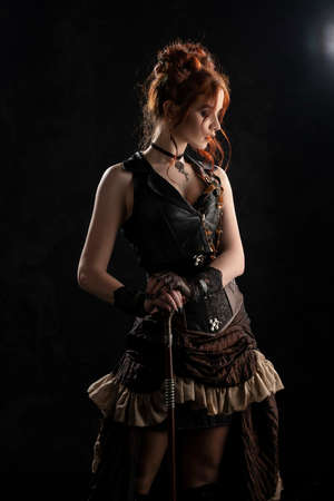 A beautiful red-haired cosplayer girl wearing a Victorian-style steampunk costume with a big breast in a deep neckline stands thoughtfully, leaning on a cane, on a dark background. Copy space.の写真素材