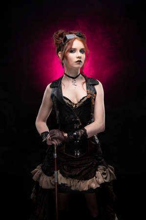 A beautiful red-haired cosplayer girl wearing a Victorian-style steampunk costume with large breasts in a deep neckline and glasses holding a cane on a black and purple background. Copy space.の写真素材