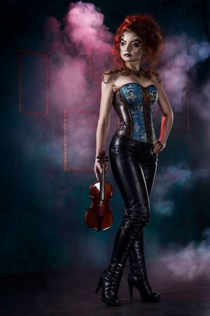 Beautiful redhead cosplayer girl wearing a steampunk corset and leather pants with a big breast holds violin in her hand in theatrical smoke. Copy space.の写真素材