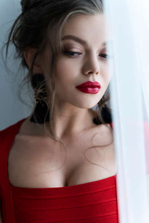 Beautiful young sexy elegant girl with big breasts, wearing a red tight dress and earrings sensually standing by the curtains at the window. Commercial and advertising design. Copy space.の写真素材