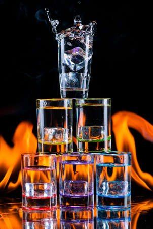 Multicolored glasses filled with alcoholic drinks, with splases of ice cubes falling inside, standing on the mirror surface on background of fire flames. On black. Conceptual design. Closeup.の写真素材