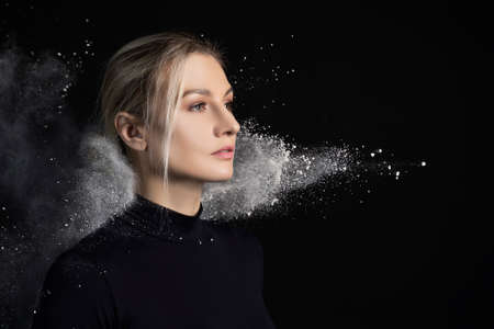Beautiful slim girl wearing a black gymnastic bodysuit covered with clouds of the flying white powder poses on a dark background. Artistic conceptual and advertising photo. Copy space.の写真素材