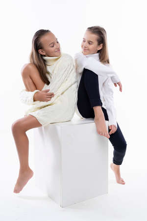 Two cute teenage girlfriends schoolgirls wearing white turtleneck sweaters, smiling sit back to back. Isolated on white. Fashion and advertising design. Copy space.の写真素材