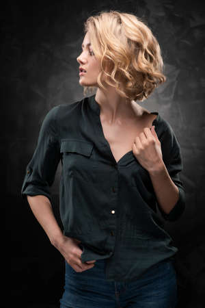 Beautiful young blonde girl with disheveled hairstyle and nude makeup, wearing a shirt and jeans emotionally posing on a gray background. She with pleasure pulls to side the shirt collar. Copy space.の写真素材