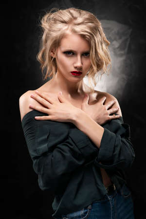 Beautiful young braless slim blonde girl with disheveled hair and aggressive makeup, wearing an unbuttoned shirt and jeans emotionally posing on a gray background. Commercial design. Copy space.の写真素材