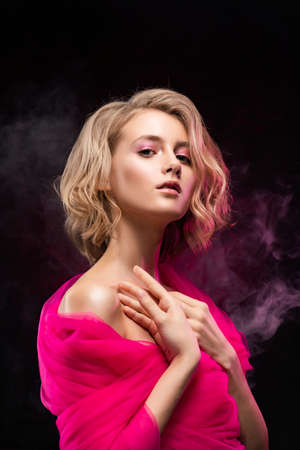 Beautiful young blonde girl with naked oily shoulders and curly hair, wrapped in a pink veil posing on a pink gradient background in theatrical smoke. Clean, healthy skin. Close-up. Copy spaceの写真素材