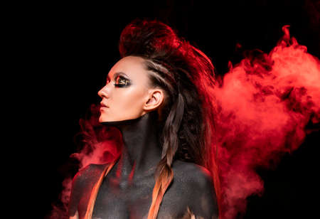 Portrait of a young savage girl in red smoke. Naked shoulders and neck are covered with black paint. Conceptual makeup with gold leaf on her eyelids. Black background. Commercial design. Copy space.の写真素材