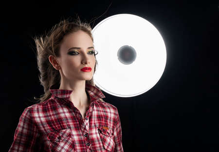 Beautiful girl model with red lips make-up, and hair flowing from the wind, wearing a casual plaid shirt near a round light on a black background. Advertising and fashion design. Copy space.の写真素材