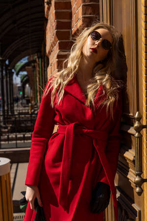 A beautiful blond girl wearing a red coat, sunglasses and gloves is standing at a brick wall lit by the bright sun. Fashionable, commercial, advertising design. Copy space.の写真素材