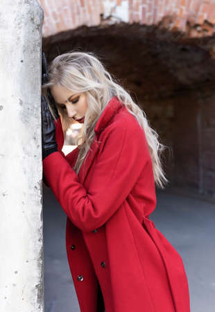 A beautiful blond girl wearing a red coat and gloves stands by a ruined wall and brick arch near an old building. Fashionable, commercial, advertising design. Copy space.の写真素材