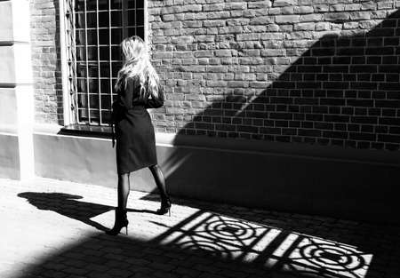 A beautiful blond girl wearing a red coat and gloves stands near the shadow of a wrought fence on the sidewalk. Fashionable, commercial, conceptual, artistic black and white design. Copy spaceの写真素材